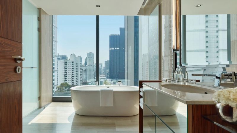 Luxury hotel bathroom with freestanding bathtub and panoramic city skyline view through floor-to-ceiling windows.